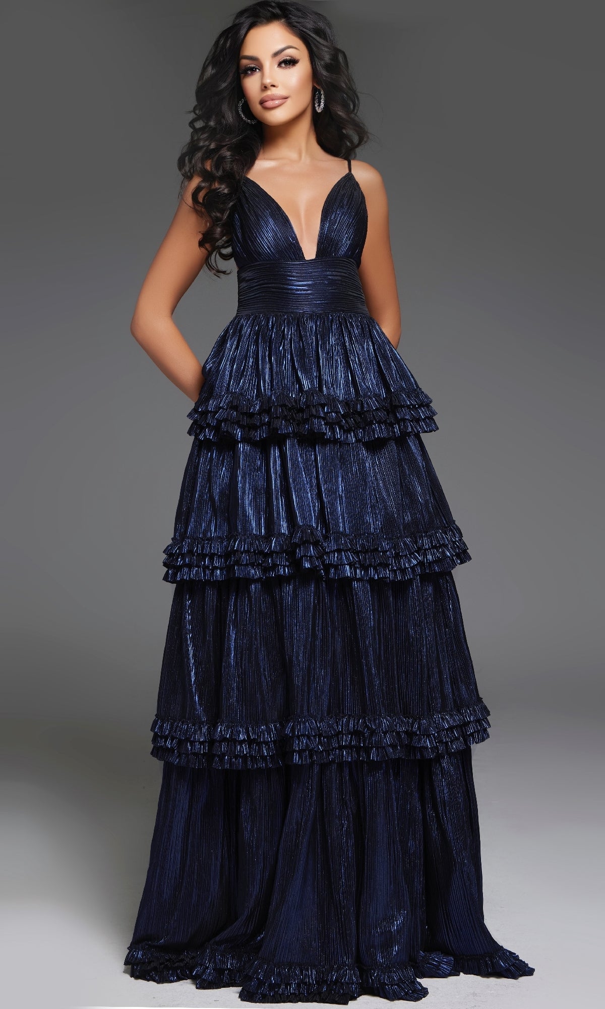 A woman stands against a plain gray background wearing the Jovani 38296, a long navy prom dress with a sleeveless, deep V-neck design and tiered ruffle details.